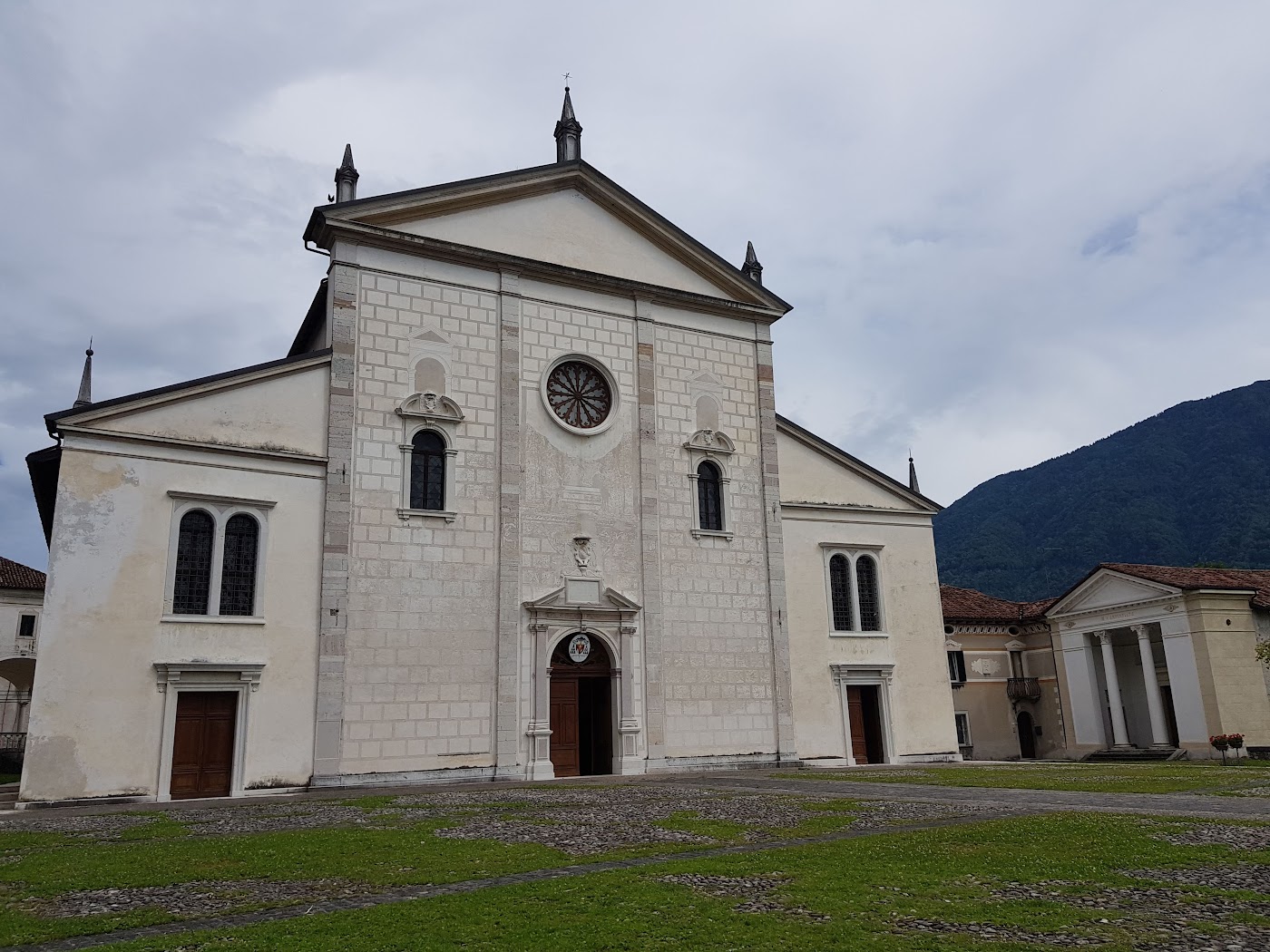 Facciata chiara di una chiesa con rosone e frontone, con le montagne sullo sfondo, a Feltre