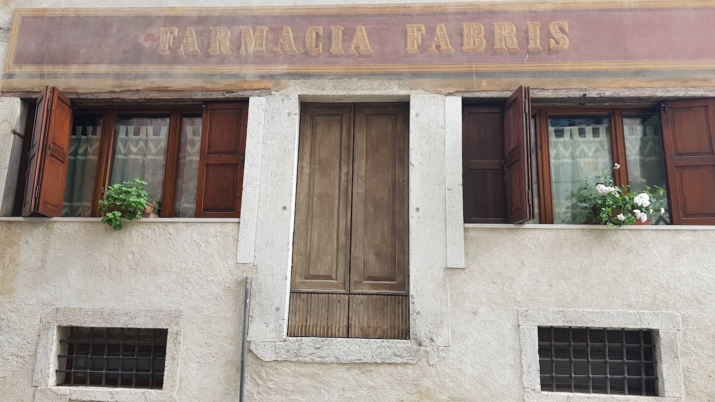 Facciata con l'insegna dipinta Farmacia Fabris, persiane in legno e fiori alle finestre